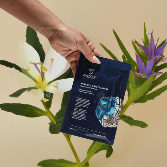 Hand holding a packet of Roccoco Midnight Crystal Sheet Mask on tan background with botanicals